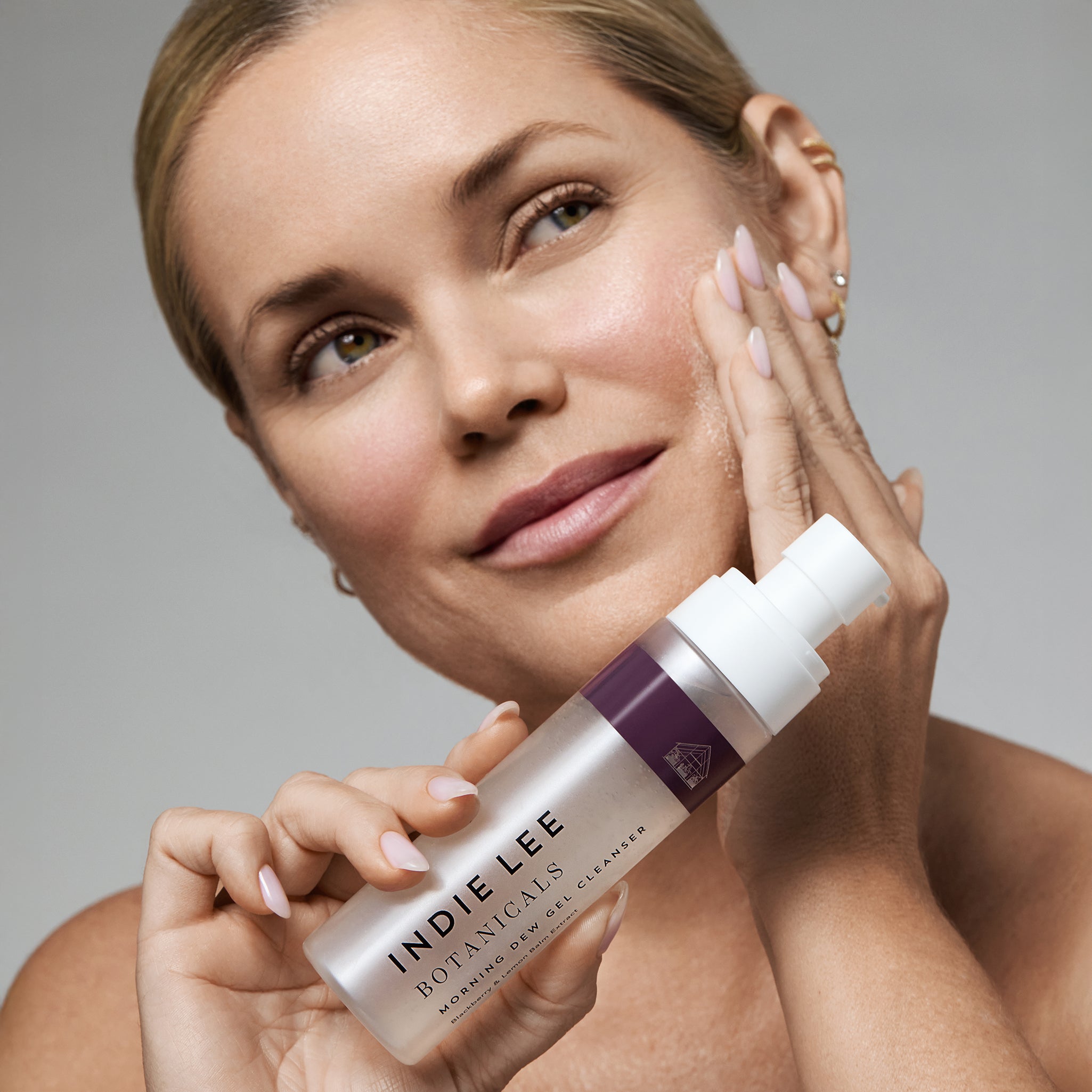 Woman holding a bottle of Indie Lee Botanicals skincare product against a neutral background