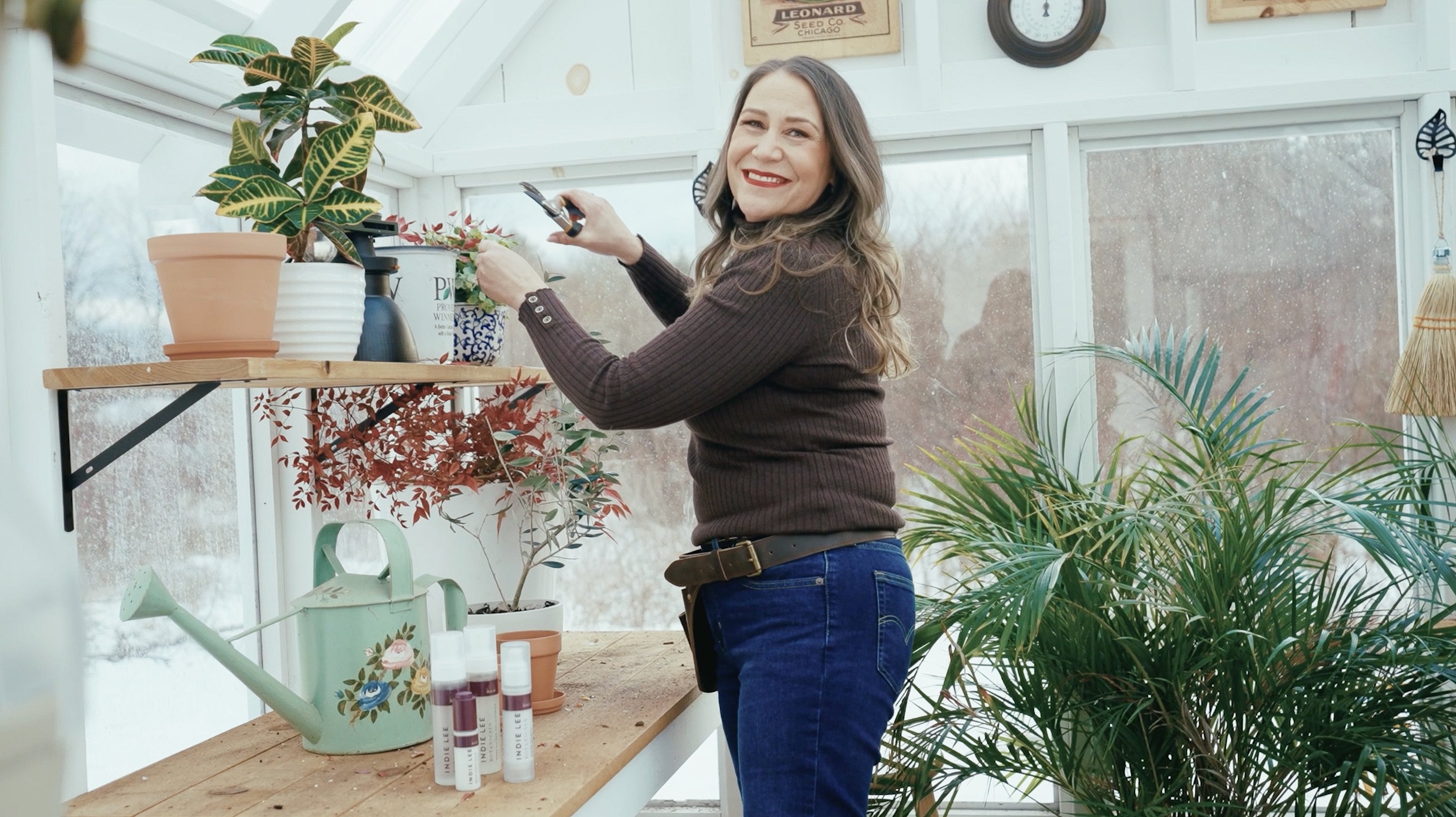 Indie Lee smiling in a greenhouse with plants and gardening tools