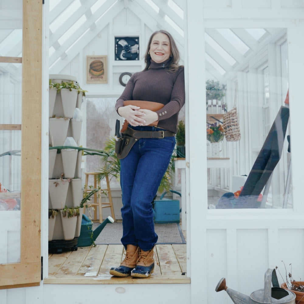 Indie Lee standing in a greenhouse holding gardening tools