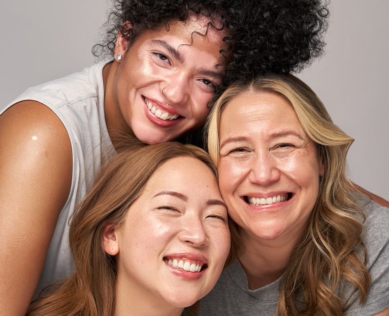 Indie Lee and two friends Yaya and Hanah posing closely together against a plain light gray studio background.
