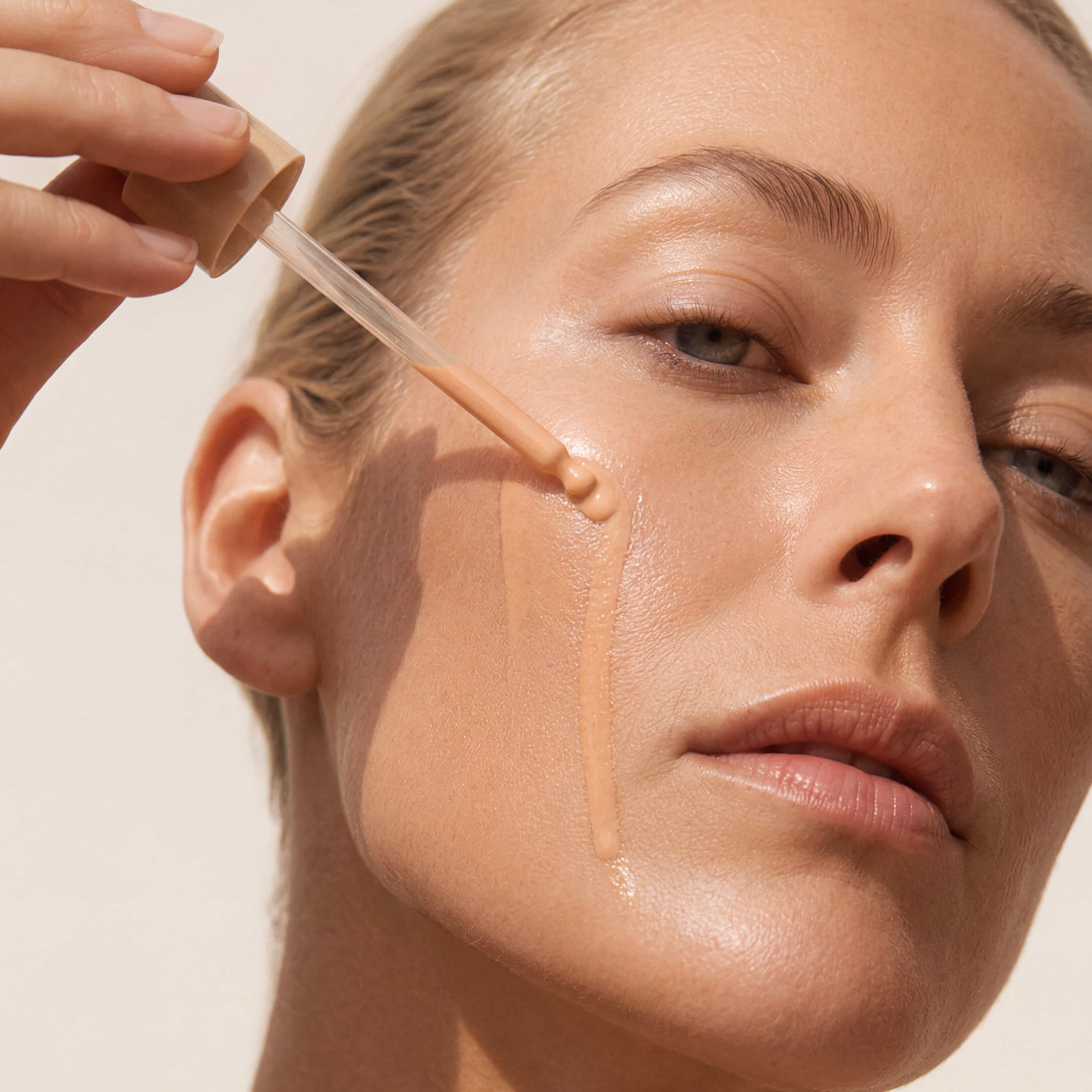 Close-up of a woman's face with a liquid makeup product being applied with a dropper against a plain light background.