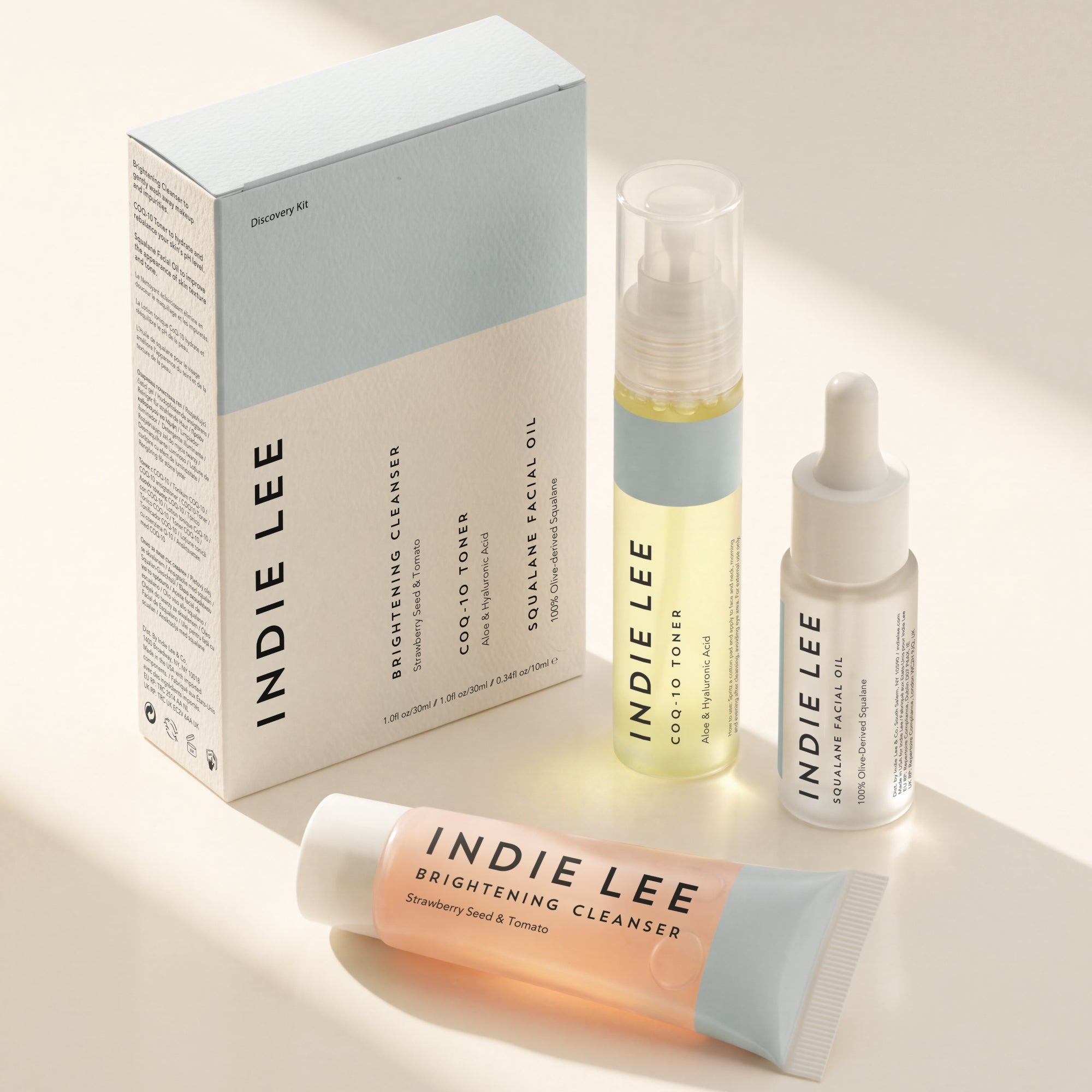 Indie Lee Discovery Kit on a warm beige background with soft shadows. The set includes a brightening cleanser tube, a cold-pressed facial oil bottle, a dropper serum bottle, and the product box.