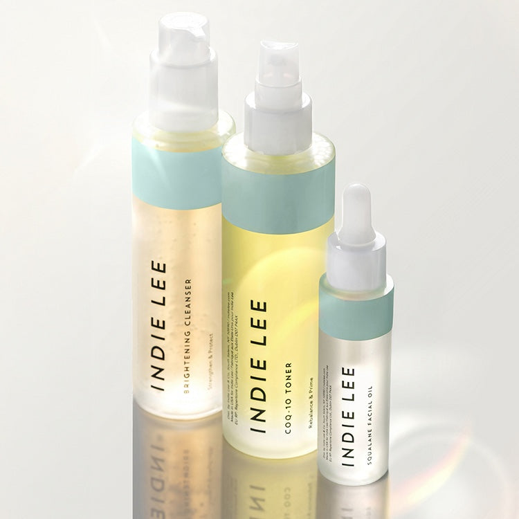 Three Indie Lee skincare bottles arranged together on a reflective white surface against a bright white background. 