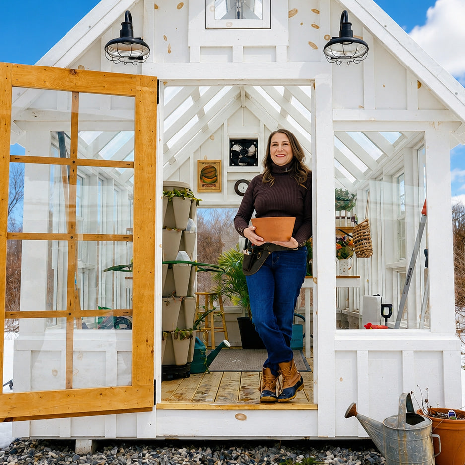 Indie Lee standing in a greenhouse holding a pot, with a white building and blue sky in the background.