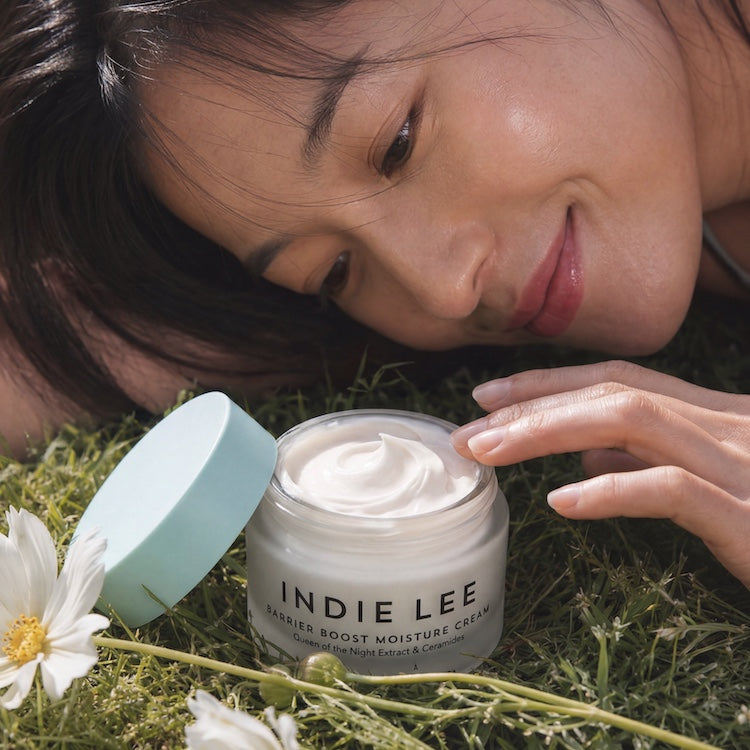 A woman lying in grass beside an open jar of Indie Lee Barrier Boost Moisture Cream, with the light blue lid resting nearby and white flowers in the foreground.