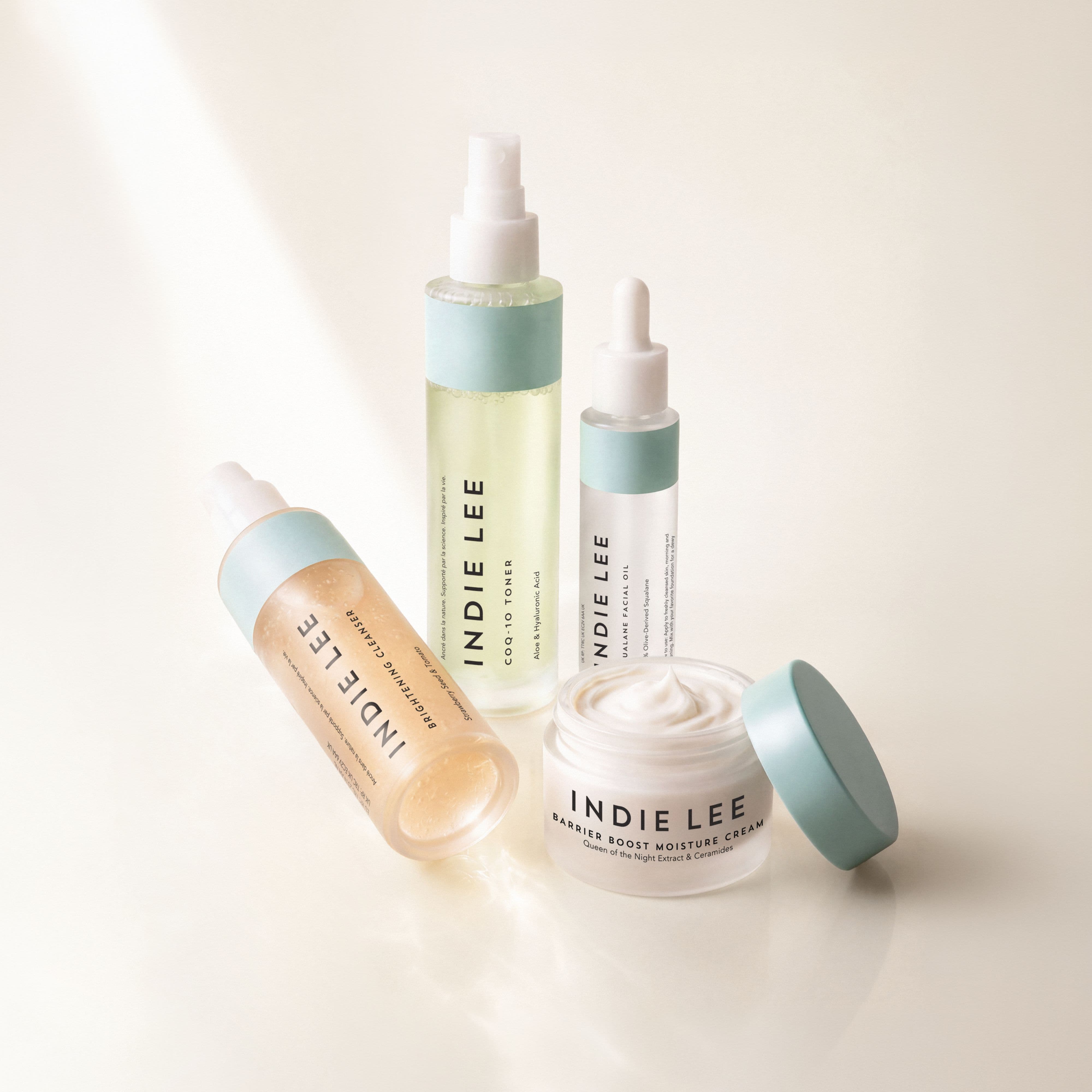 Four Indie Lee skincare bottles and jars arranged on a clean white surface with soft sunlight and shadows.