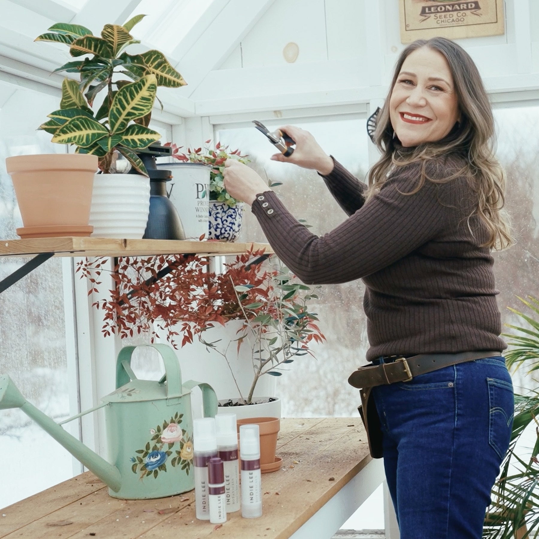 Indie Lee smiling inside her greenhouse with plants and gardening tools.