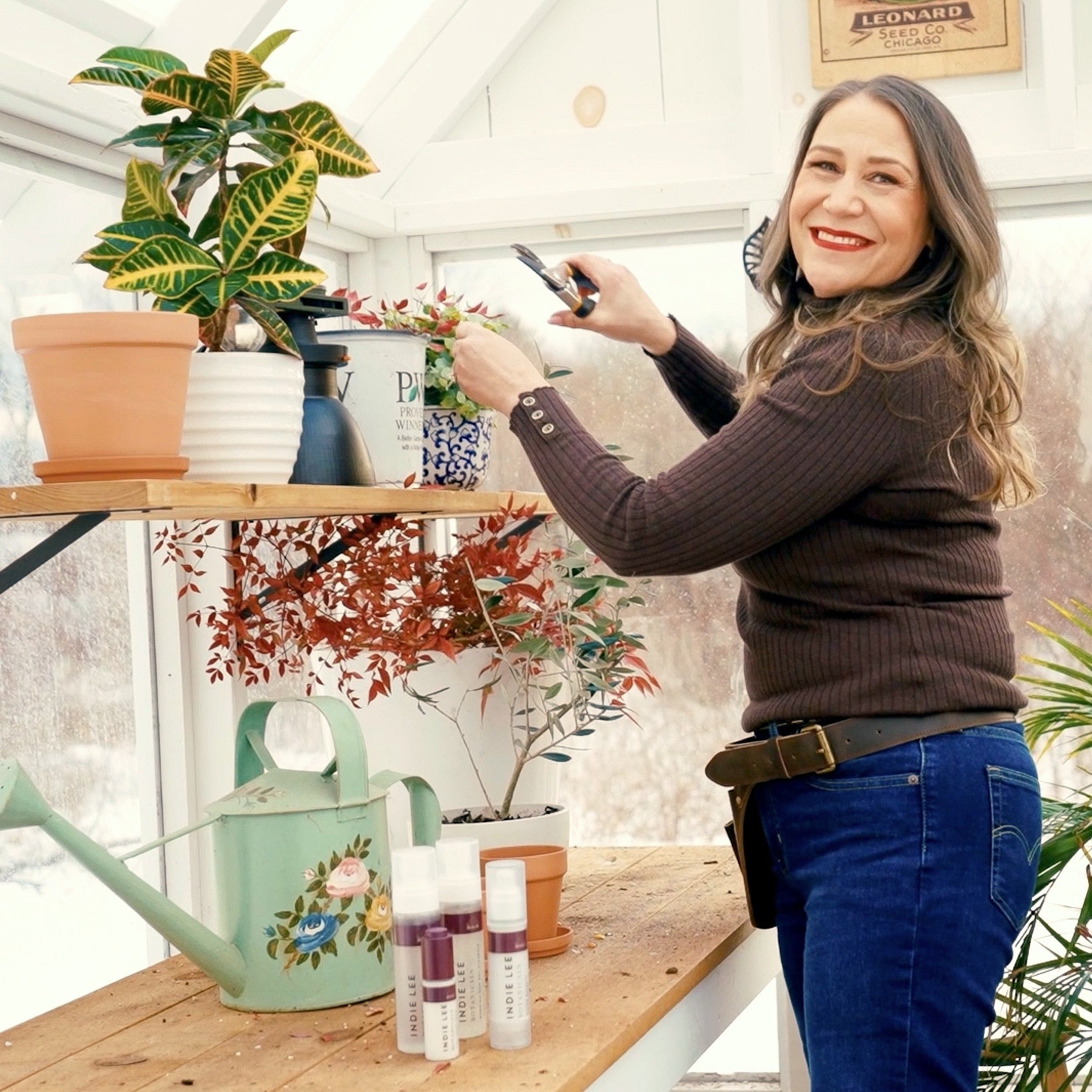 Indie Lee in her greenhouse standing near potted plants on wooden shelves.