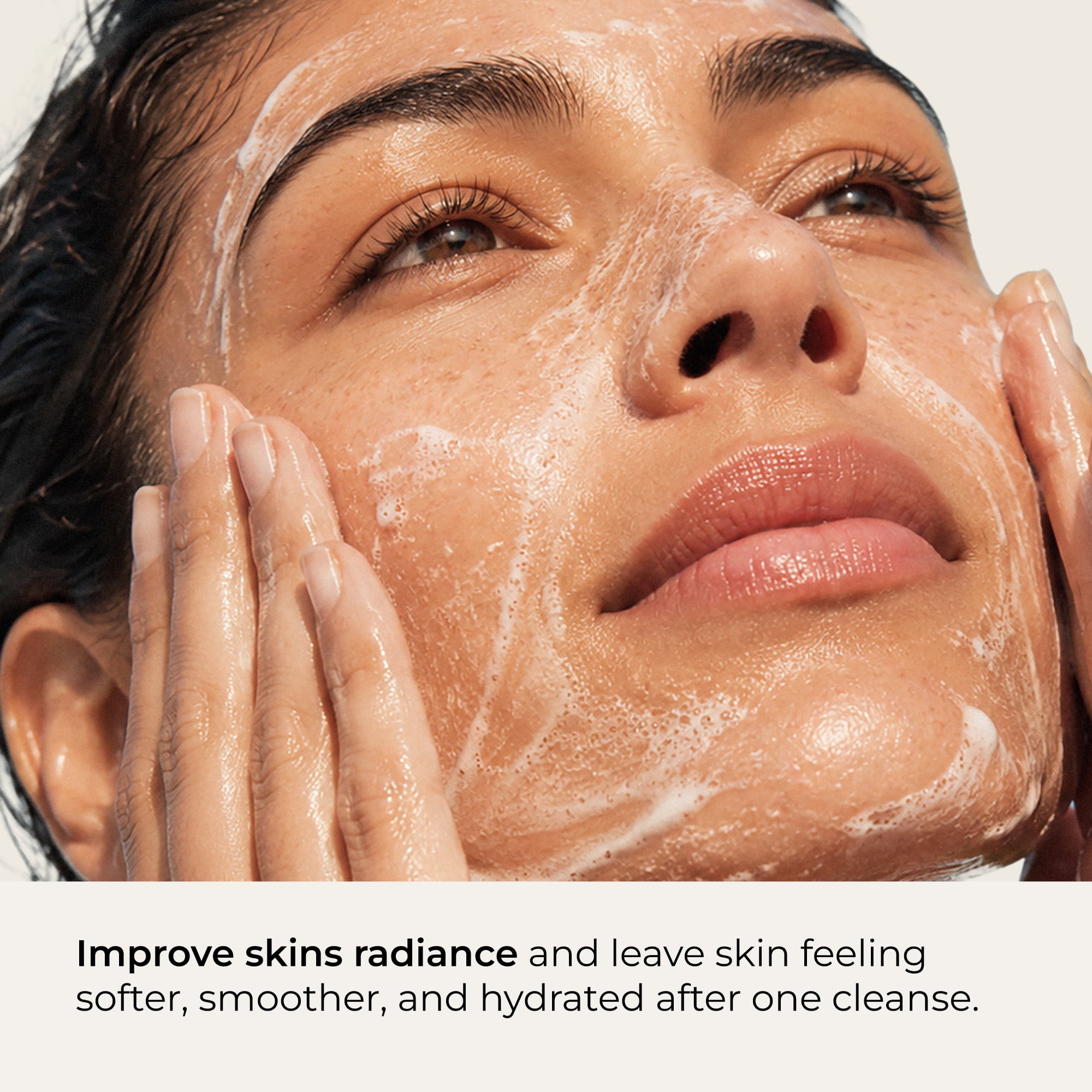 Close-up of a woman's face with cleanser lathered across it with text at the bottom reading 'Improve skin's radiance and leave skin feeling softer, smoother, and hydrated after one cleanse.'