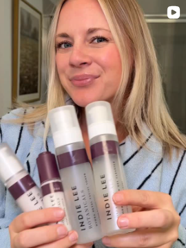 Woman holding multiple bottles of Indie Lee skincare products in a room with a painting on the wall.