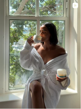 Woman in a white robe sitting by a window, holding a cup and a jar of Indie Lee skincare product.